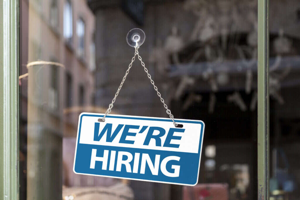 IT recruitment has never been harder. Close-up on a blue and white sign in a window with written in "We're hiring".
